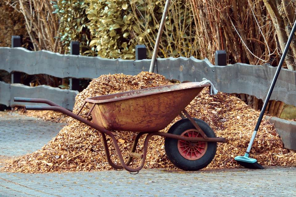 Empty wheelbarrow next to a pile of mulch, a fence and a broom