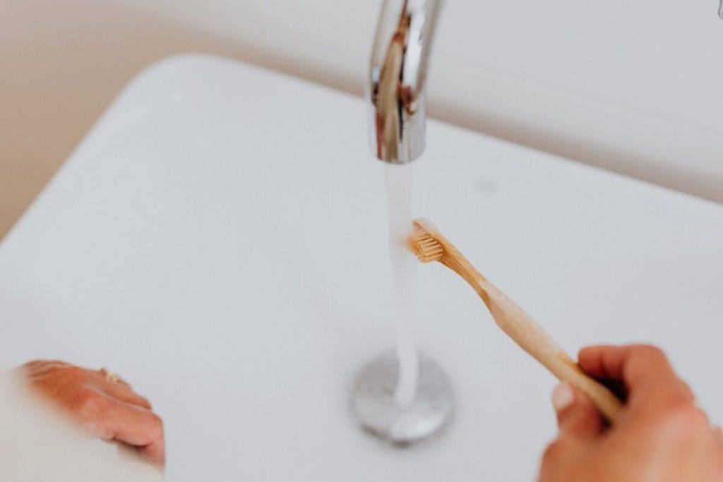 A person using a bamboo toothbrush in a sink bathroom with flowing water.
